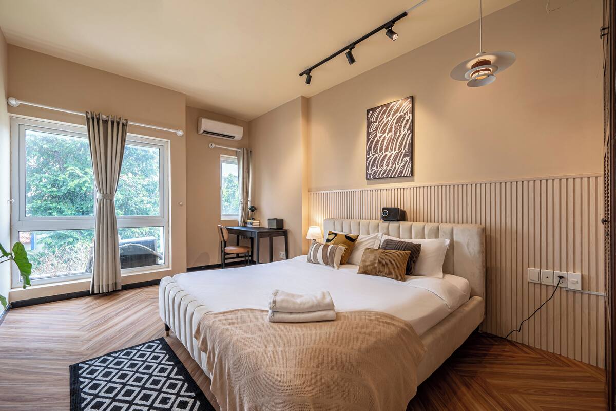 A hotel room with a large bed, a desk with a chair, a window with curtains, and a radiator on the wall.