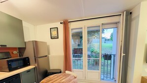Eine kompakte Küche mit Mikrowelle, Kühlschrank und Geschirrspüler. Ein Holztisch mit Stühlen, ein gemustertes Teppich und eine Glastür, die auf einen Balkon mit Blick auf den Garten führt.