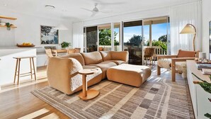 A spacious living area with a large sectional sofa, a small round table, and a dining table with chairs. There is a large window providing a view of the outdoors.