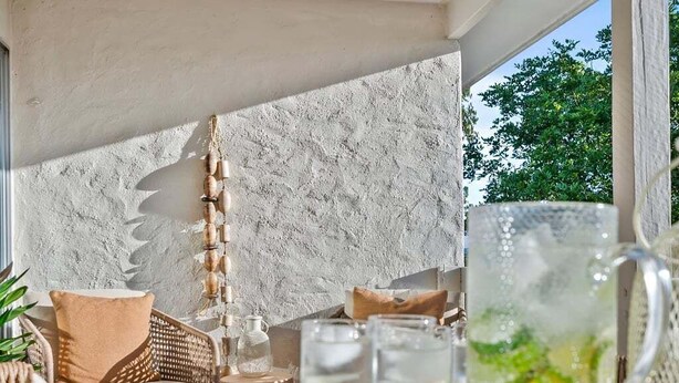 A cozy outdoor seating area with a wicker sofa, a wooden table with drinks, and a textured wall.