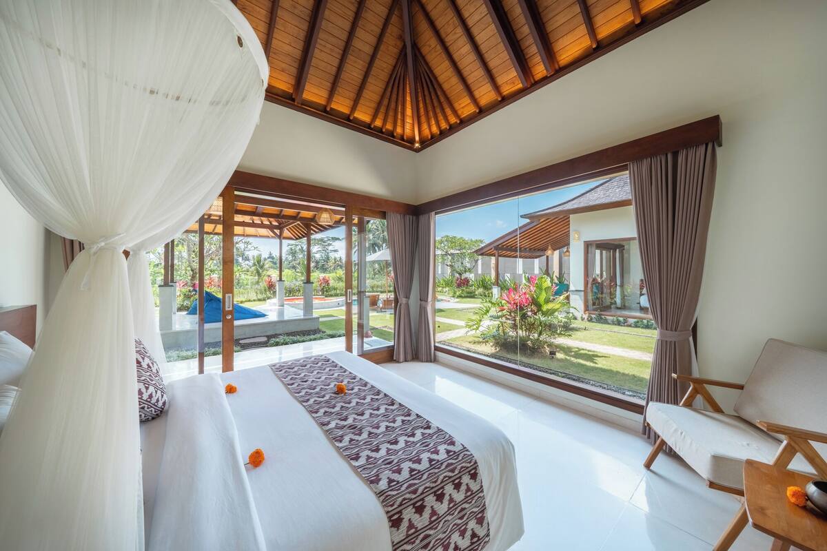 A bedroom with a large bed, a canopy, and a view of a garden and pool.