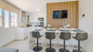 A modern kitchen with a central island, bar stools, and a flat-screen TV mounted on a wooden panel.