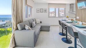 A modern living room with a grey sofa, a dining area with bar stools, and a kitchen with a view of the sea.