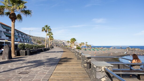 A coastal walkway with palm trees, a beach, and a hotel in the background.