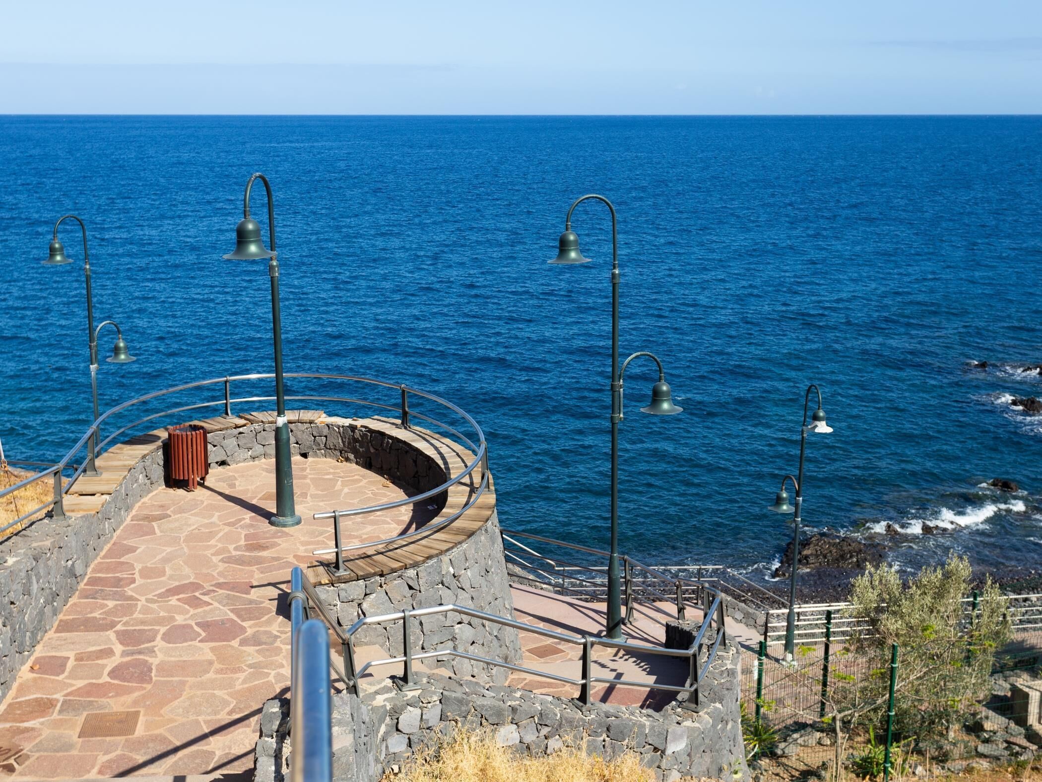 A stone-paved circular viewing platform with metal railings and a stone wall, overlooking a blue sea.