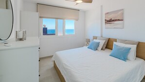 A bedroom with a bed, a ceiling fan, a window with a view of the sea, and a painting on the wall.