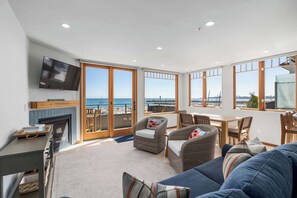 A spacious living room with a fireplace, comfortable seating, a television, and large windows offering a view of the sea.