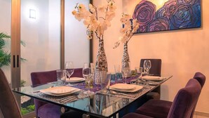 Uma sala de jantar com mesa de vidro, cadeiras roxas, pratos brancos e copos de vidro.