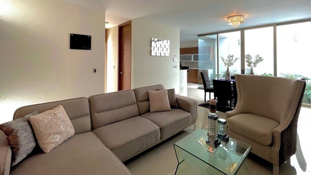 Uma sala de estar moderna com sofá, poltrona e mesa de centro de vidro. Vê-se um candeeiro, uma televisão de ecrã plano e um espaço de refeições ao fundo.