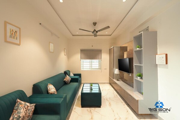 Una sala de estar moderna con un sofá verde, una mesa de centro de vidrio, un ventilador de techo y un televisor montado en un mueble.