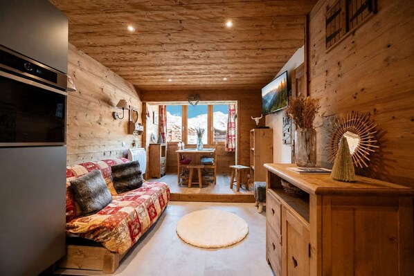 Interior acogedor de una cabaña con detalles de madera, un sofá con cojines estampados, un comedor y vistas a montañas a través de las ventanas.