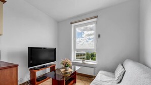 En moderne stue med et fladskærms-tv på et træstel, et glas sofabord med en vase blomster, en grå sofa og et stort vindue med udsigt til bygninger og træer.