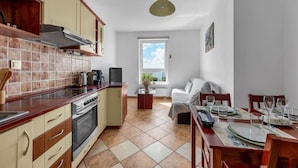 Et kompakt køkken med komfur, ovn og moderne hårde hvidevarer. Der er et spiseområde med et bord dækket til to, og et vindue med udsigt til det fri.