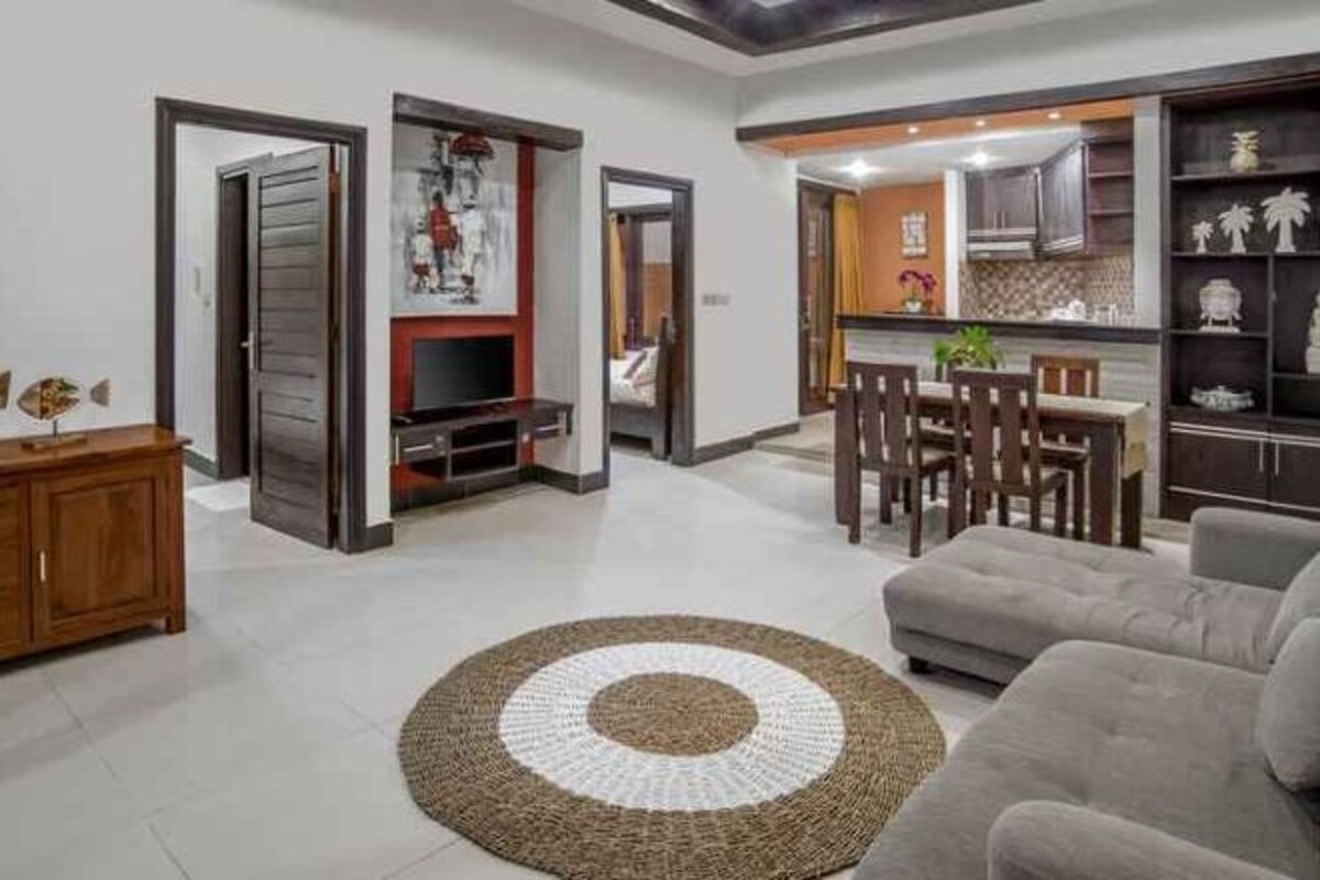 Una sala de estar con un sofá, una mesa de centro y una alfombra. Hay una zona de cocina con armarios y una mesa de comedor.