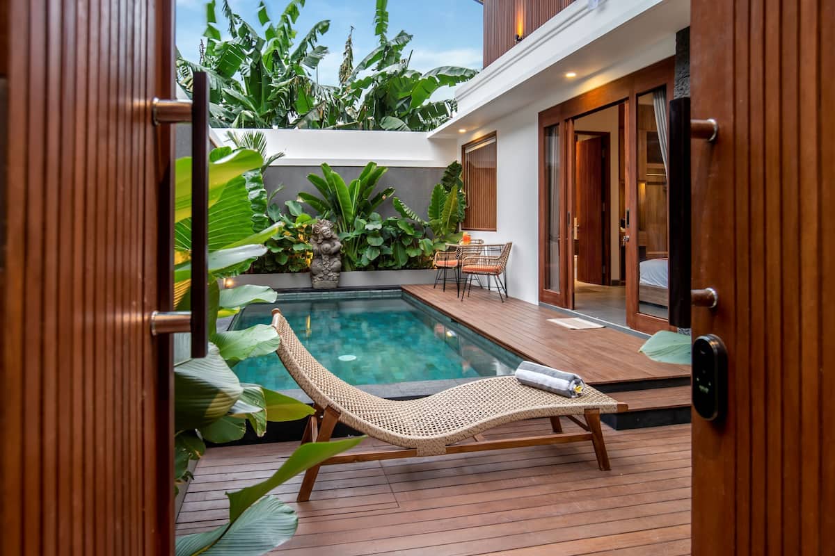 A modern outdoor pool area with a lounge chair, wooden deck, and lush tropical plants.