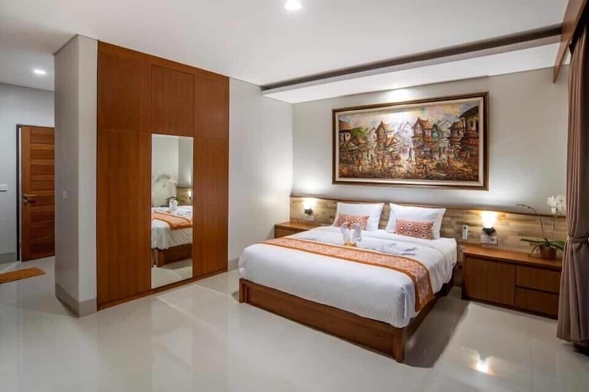 A modern bedroom with a large bed, wooden wardrobe, a painting on the wall, and a nightstand with a lamp.