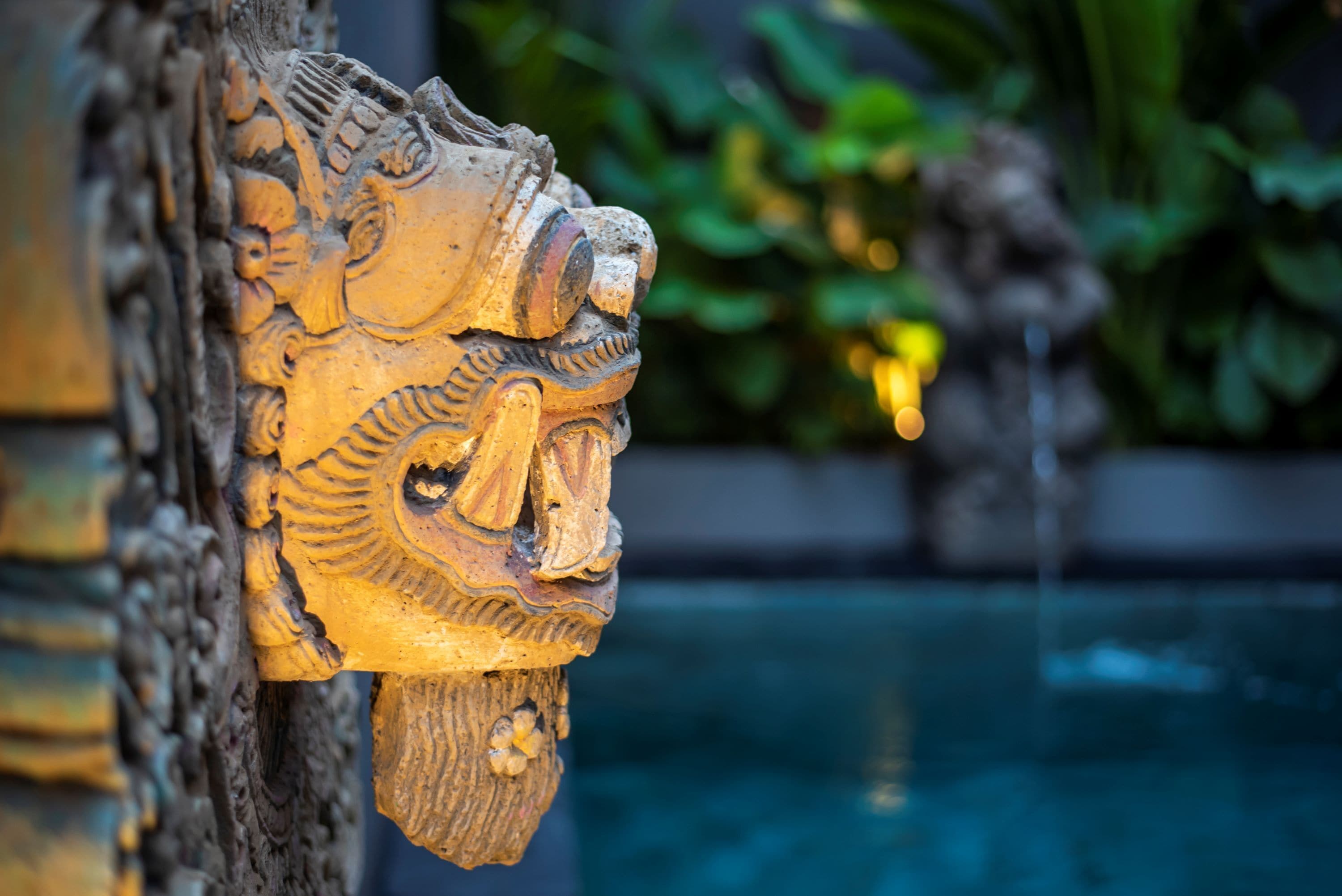 A detailed carved wooden head with a flowing beard, set against a blurred background of greenery and water.