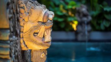 A detailed carved wooden head with a flowing beard, set against a blurred background of greenery and water.