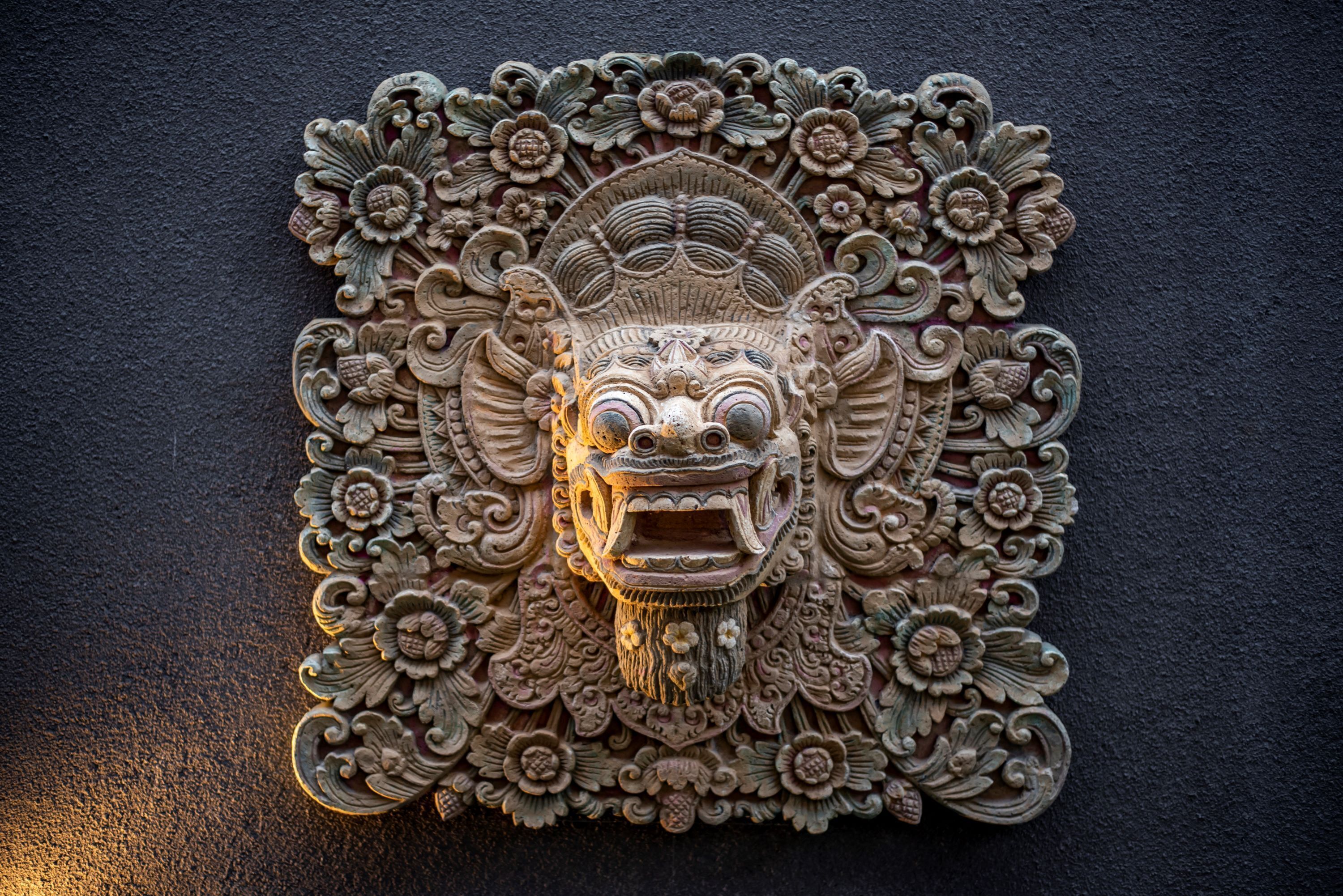 Eine detaillierte Steinschnitzerei eines Gesichtes, das einem mythologischen Wesen zuzuordnen ist, umgeben von floralen und geschwungenen Ornamenten.