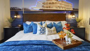 A bedroom with a large bed, nautical-themed pillows, a nightstand with a clock, and a tray with drinks and flowers.