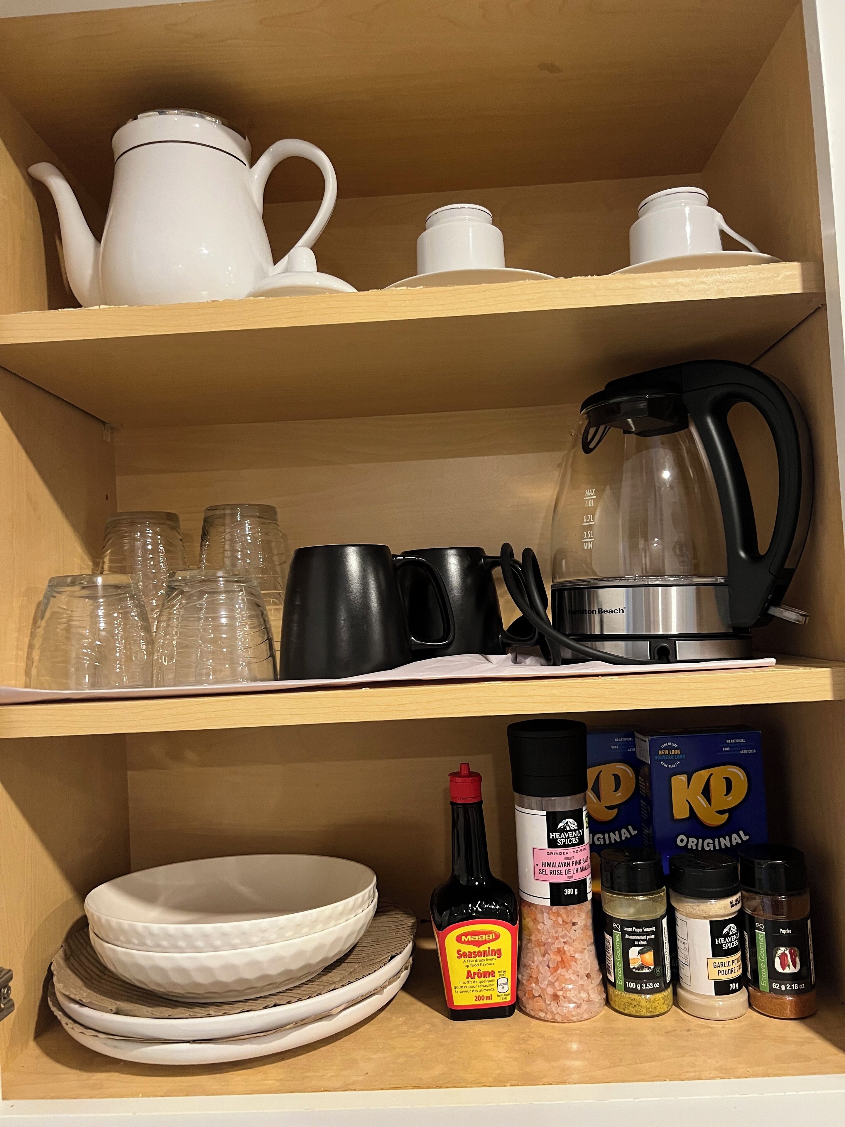 Un estante de cocina con teteras blancas, tazas negras, una cafetera de vidrio y varios recipientes de especias.