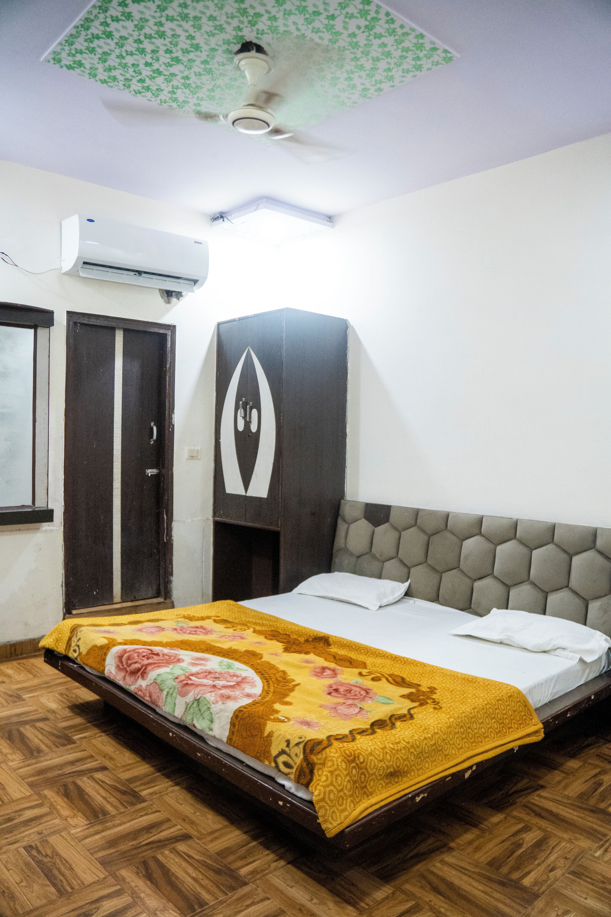 A bedroom with a wooden floor, a bed with a floral bedspread, a ceiling fan, an air conditioning unit, and a door.