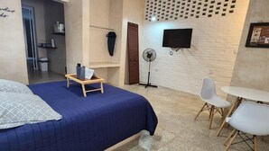 A bedroom with a bed, bedside table, fan, TV, and a small dining area with chairs.