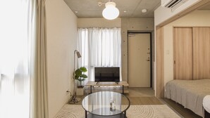 Habitación de hotel pequeña con una cama, un televisor, una mesita con superficie de vidrio y una planta en maceta.