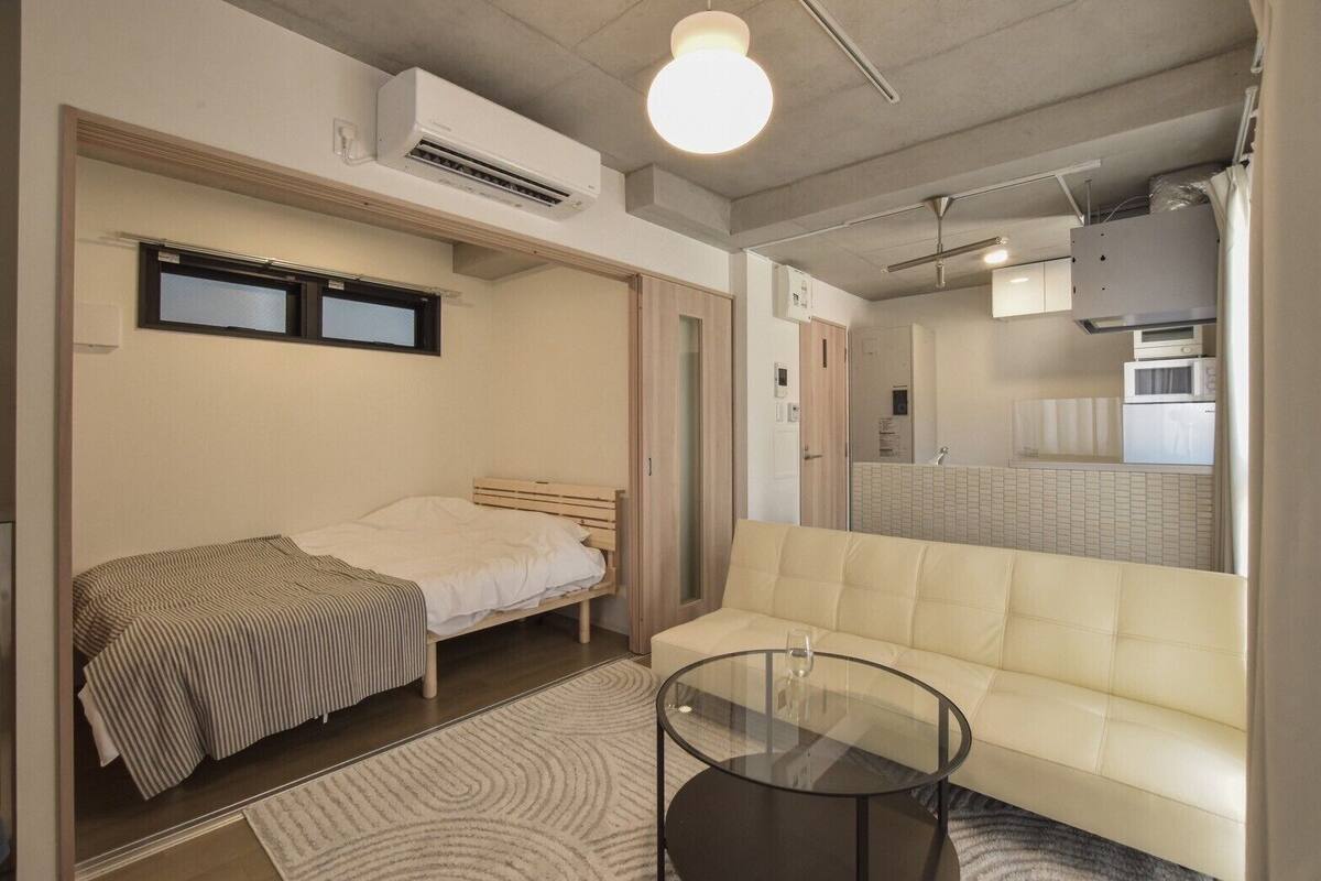 A compact bedroom with a bed, a sofa, a glass table, and a kitchenette visible through an open door.
