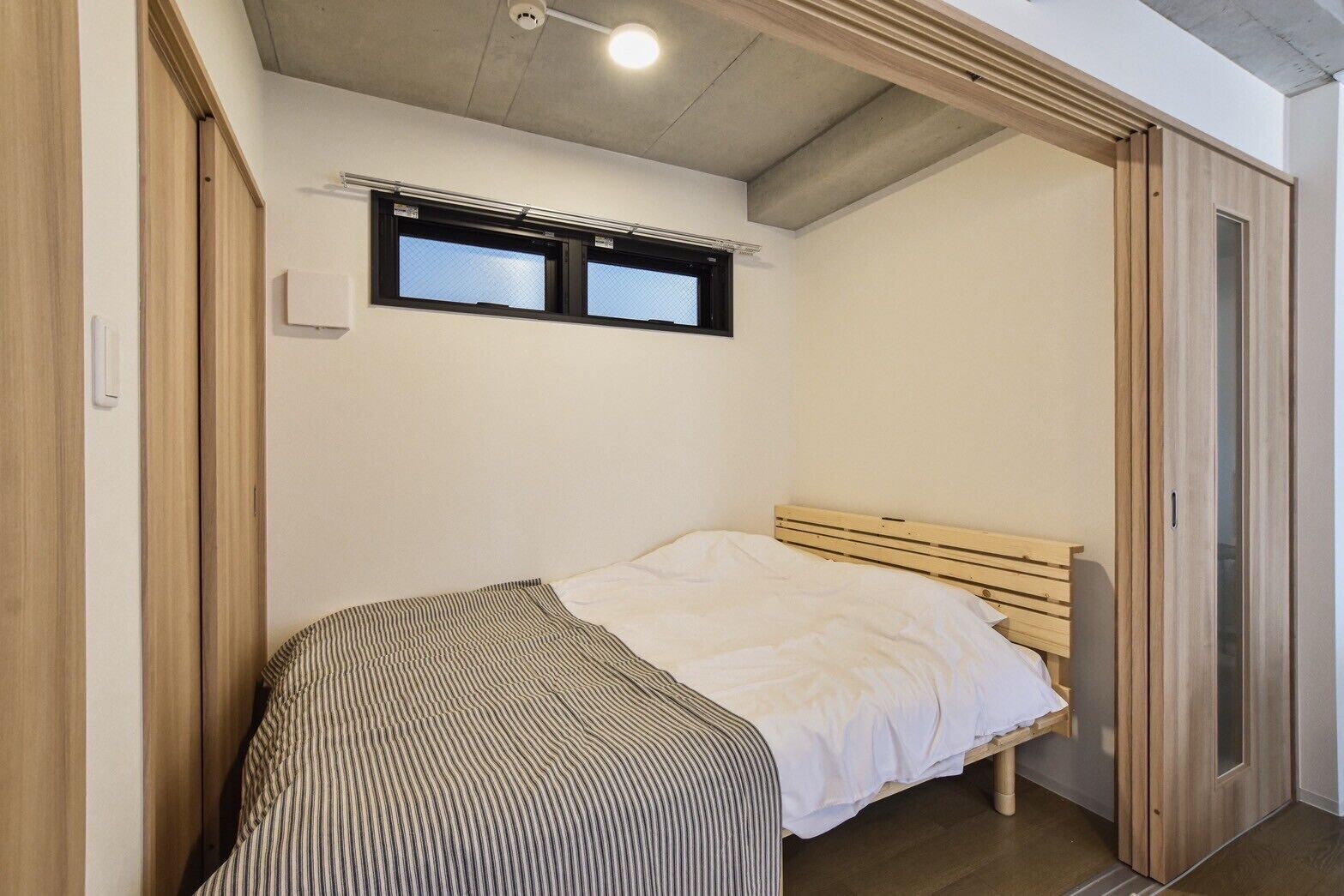 Une petite chambre avec un lit simple, une tête de lit en bois, une fenêtre située au-dessus du lit et une porte coulissante en bois sur le côté droit.