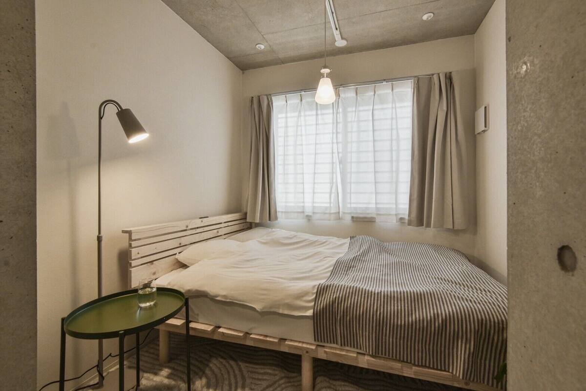 A small, single-bed room with a window, a bedside table, and a floor lamp.
