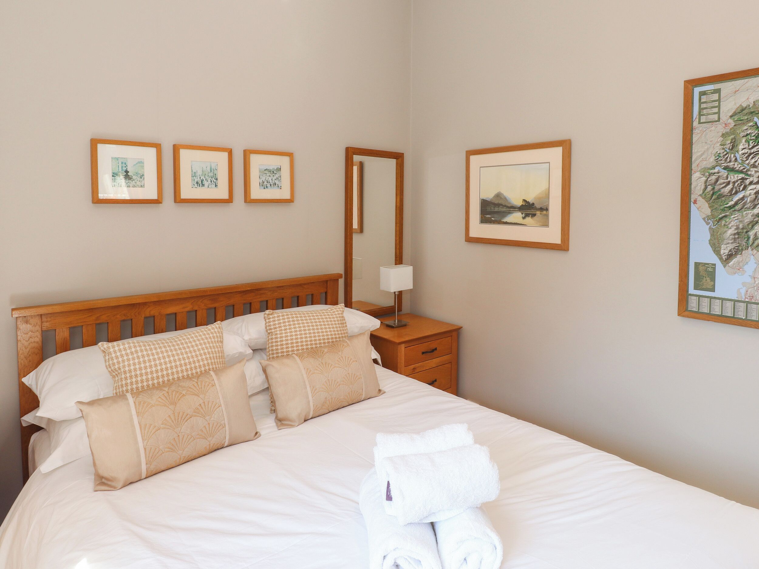 A neatly made bed with multiple pillows and a rolled towel, a wooden nightstand with a lamp, and three framed pictures on the wall.