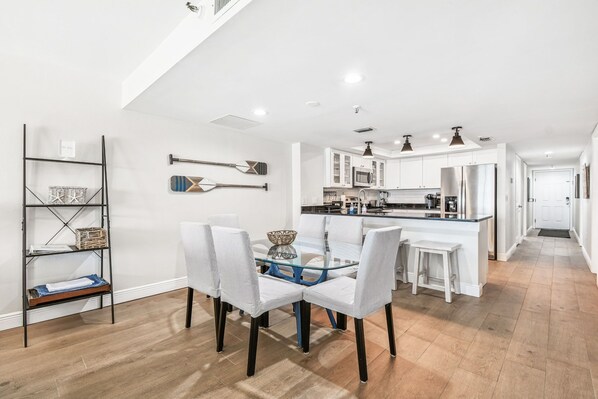 Una cocina moderna con armarios blancos, una isla central y un comedor con mesa de vidrio y sillas blancas.