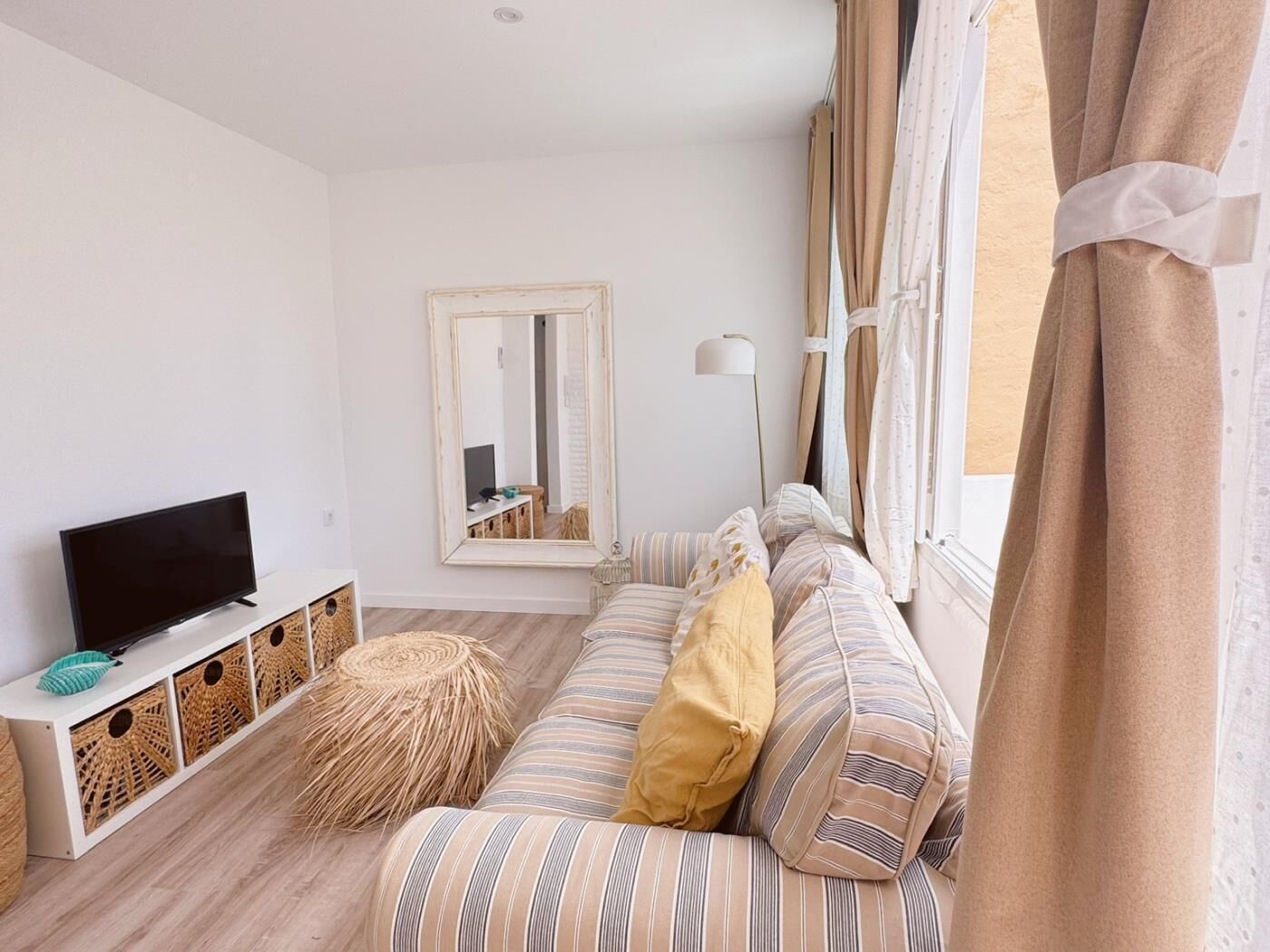 Un salon avec un téléviseur posé sur un meuble blanc, un canapé à rayures agrémenté de coussins, un grand miroir et un lampadaire.