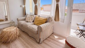 Una sala de estar acogedora con un sofá a rayas, un taburete redondo tejido y vistas de un edificio a través de una ventana con cortinas.