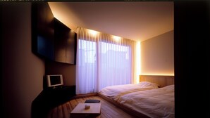 A hotel room with a bed, a bedside table with a lamp, a TV on a stand, and a window with curtains. - Linca (Zushi)
