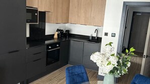 A modern kitchen with black appliances, wooden cabinets, and a dining area with blue chairs and a glass table. - Modern Newly Renovated flat - Elstree & Borehamwood Railway Station (Hertfordshire)