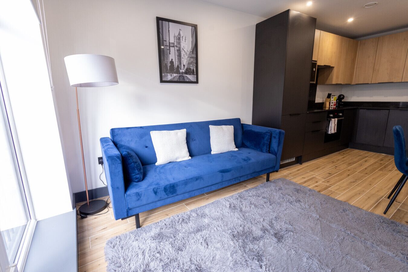 A modern living room with a blue sofa, a grey rug, and a kitchen area with wooden cabinets and a black countertop.