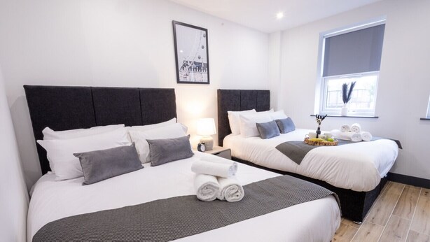 Habitación de hotel con dos camas, cada una con sábanas blancas y almohadas grises. En la pared, un cuadro enmarco; una ventana con cortina; y una mesita de noche con lámpara y una cesta de fruta.