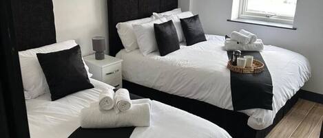 Habitación de hotel con dos camas, cada una con cabeceros negros y ropa de cama blanca. Hay una mesita de noche pequeña con una lámpara, y una ventana que proporciona luz natural.