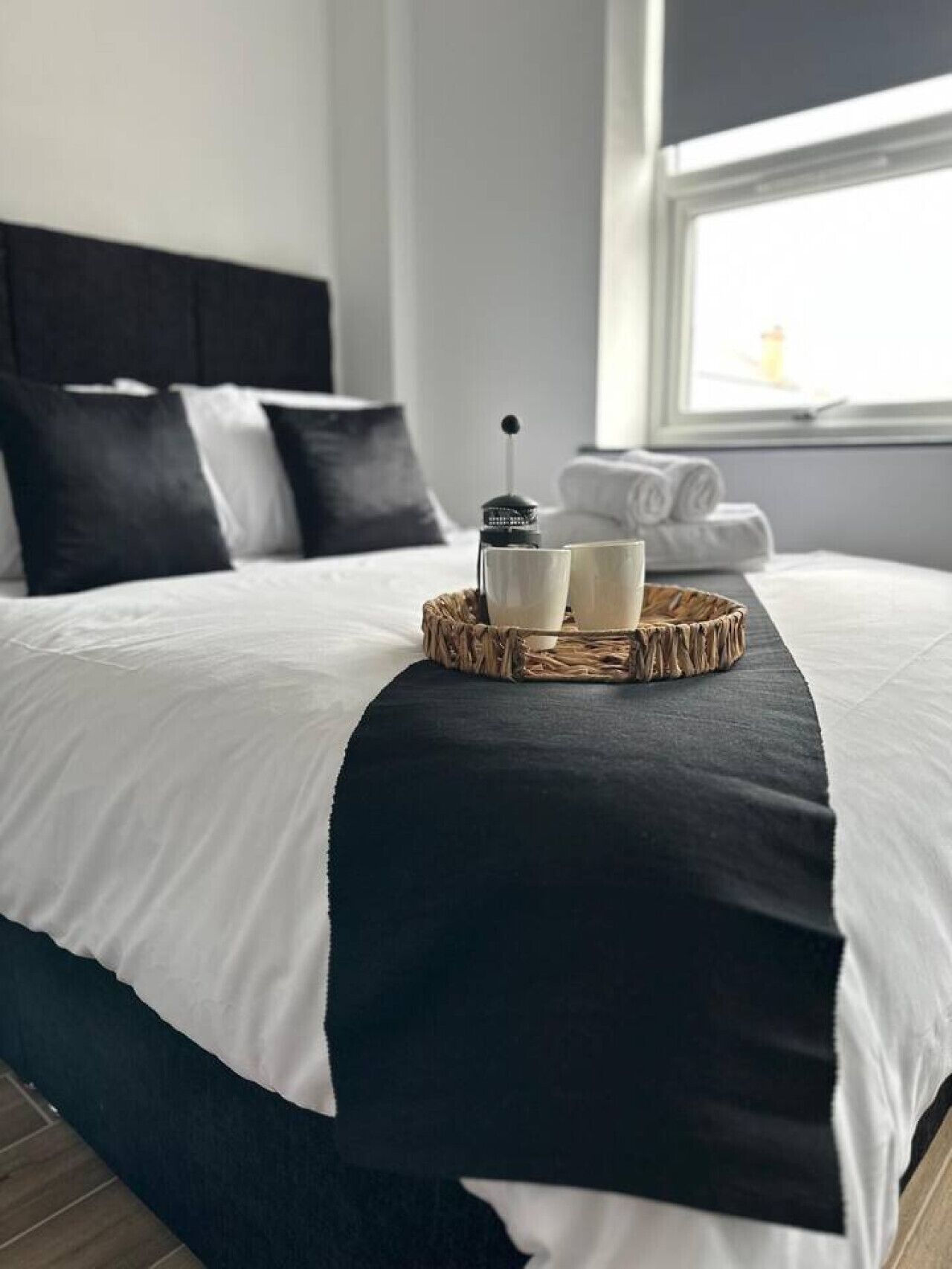 A neatly made bed with a black and white bedspread, a woven tray with two cups and a coffee maker, and towels by the window.