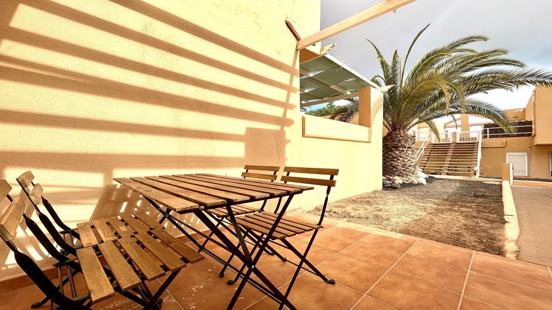 A patio with a wooden table and chairs, a palm tree, and a staircase leading to a building.