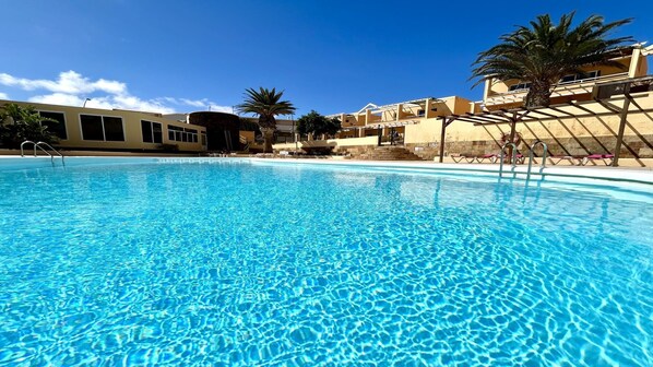 A clear blue pool with a pool ladder, surrounded by palm trees and buildings with balconies.