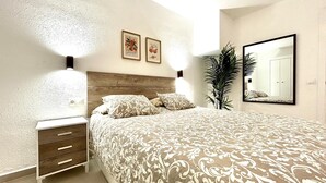Un dormitorio con cabecera de madera, una cama grande con ropa de cama estampada, una mesita de noche con cajones, dos lámparas fijadas a la pared, una planta alta y un espejo.