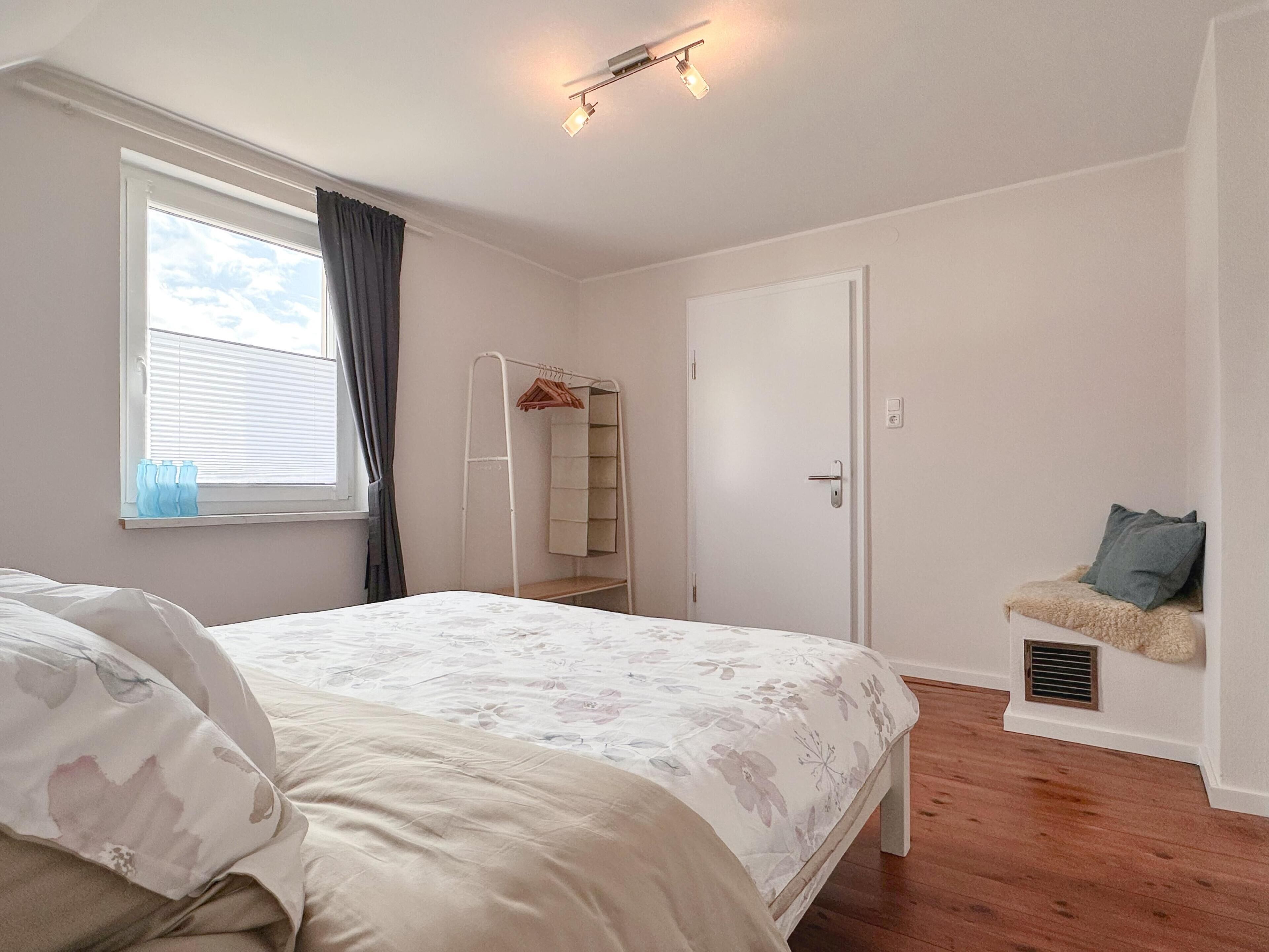 A bedroom with a bed, a wardrobe, a window with curtains, and a small seating area with a cushion.