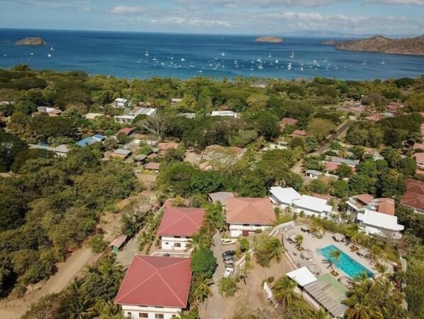 An aerial view of a coastal residential area with houses, a swimming pool, and a view of the sea with boats.