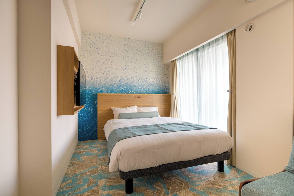 Habitación de hotel con cama, cabecera de madera, televisor montado en la pared y ventana con cortinas.