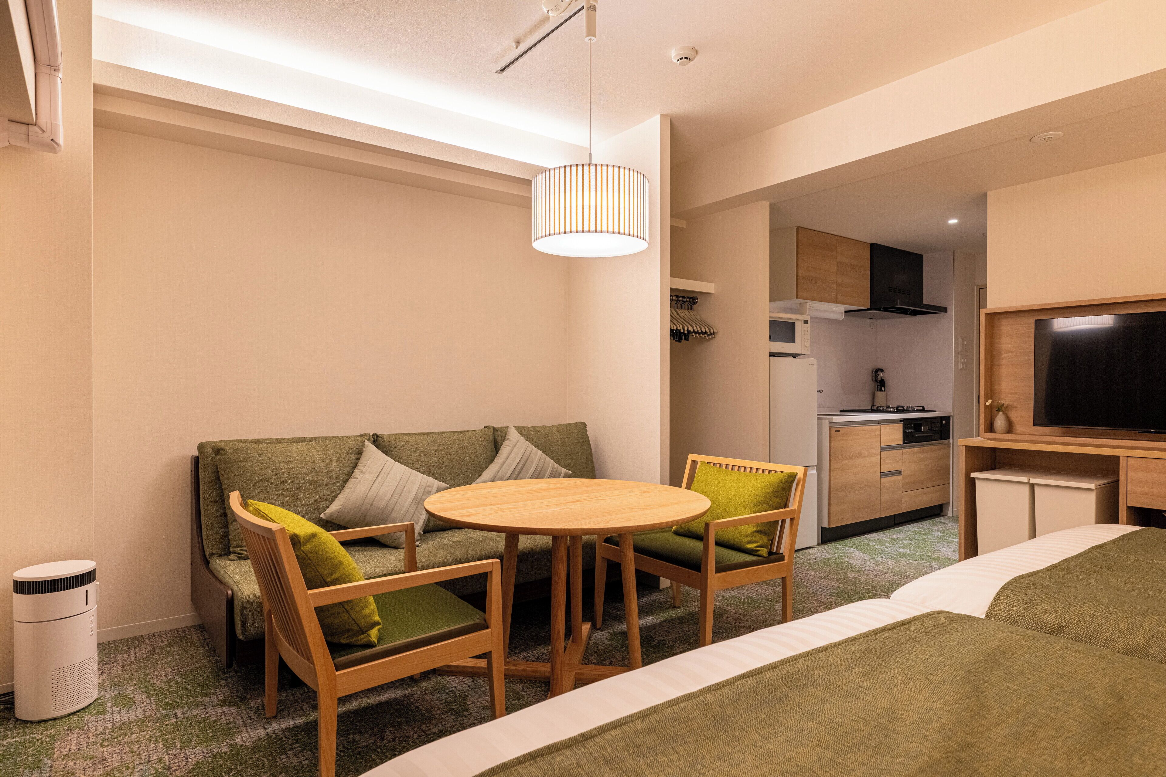 A modern hotel room with a green sofa, wooden chairs, a round table, a TV, a kitchen area, and a trash bin.