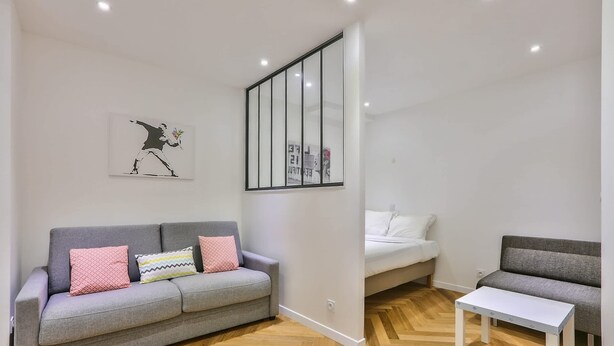 Un salon moderne avec un canapé gris, une table blanche et un lit avec une literie blanche.