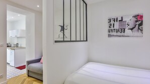 Une chambre à coucher moderne avec un lit, un fauteuil gris et, au fond, une petite cuisine.